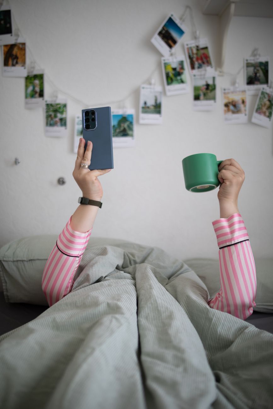 hands holding cup and cellphone over bedclothes
